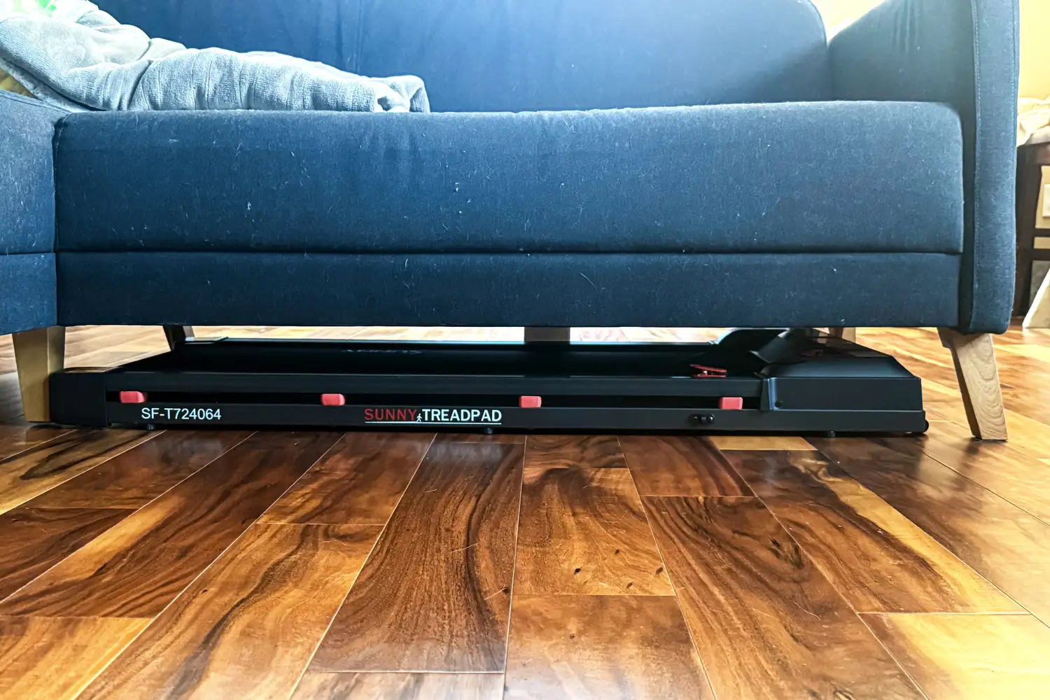 The Sunny Health & Fitness Treadpad 100 Smart Wi-Fi Connected Treadmill stored under a couch