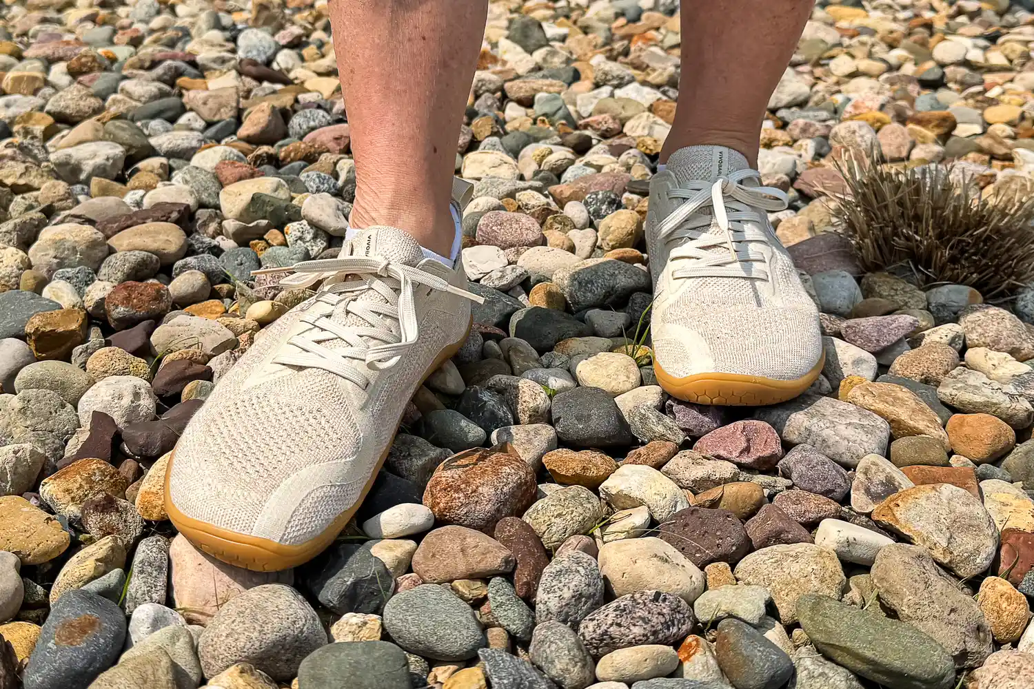 A person rocks on rocky ground while wearing the Vivobarefoot Women's Primus Lite Knit Natural Sneakers