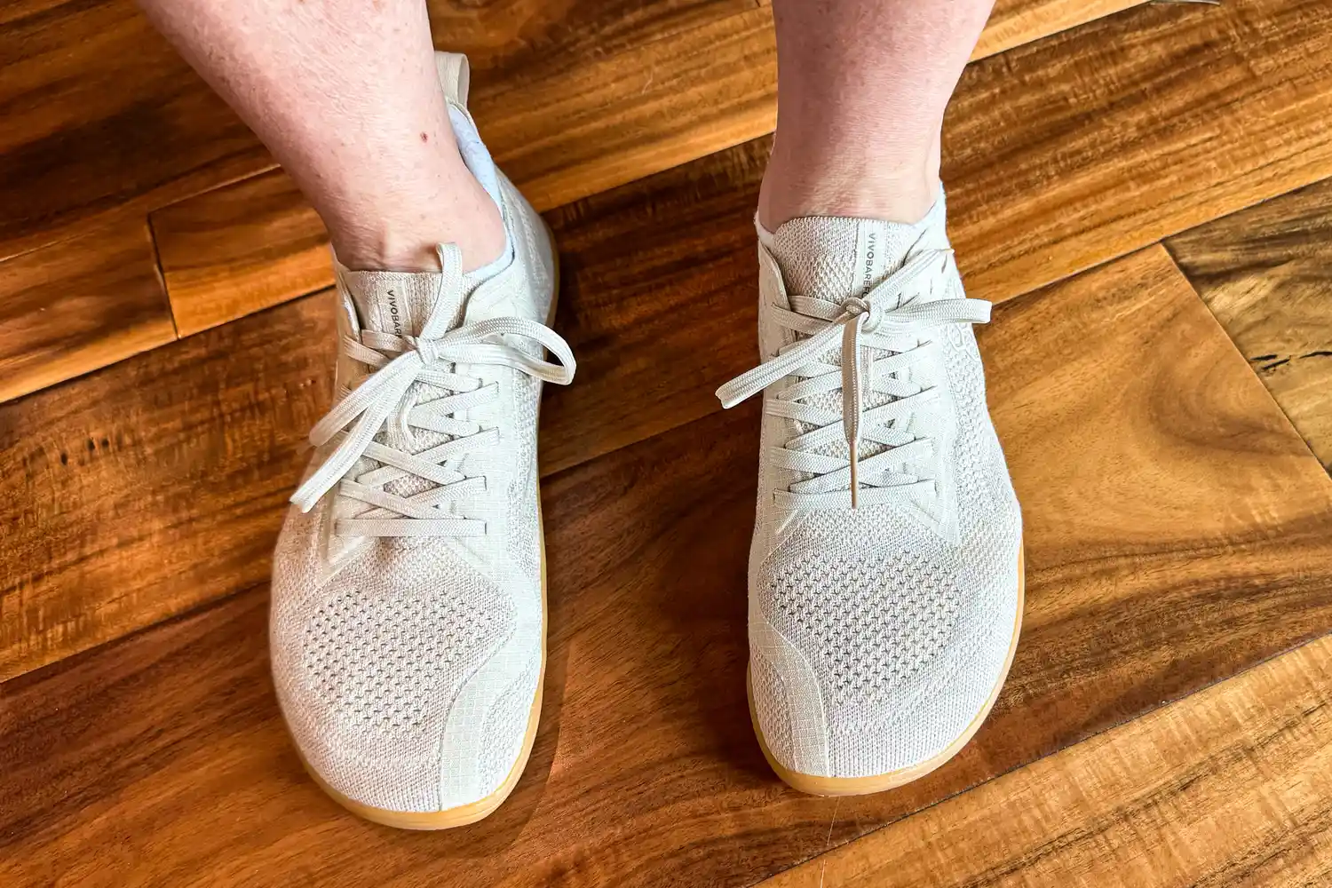 An overhead view of a person wearing the Vivobarefoot Women's Primus Lite Knit Natural Sneaker on a wooden floor