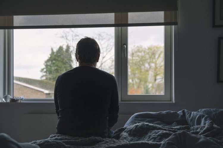 sad man sits on bed looks out window