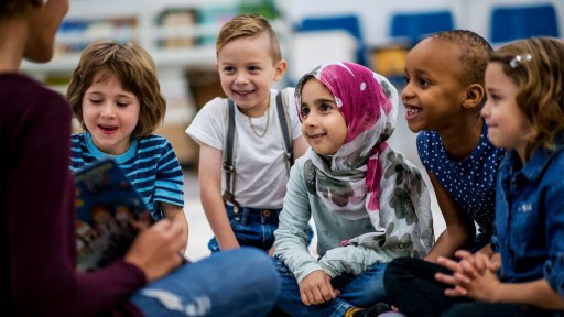 Five preschool children listen to a teacher reading a story while sitting on the floor